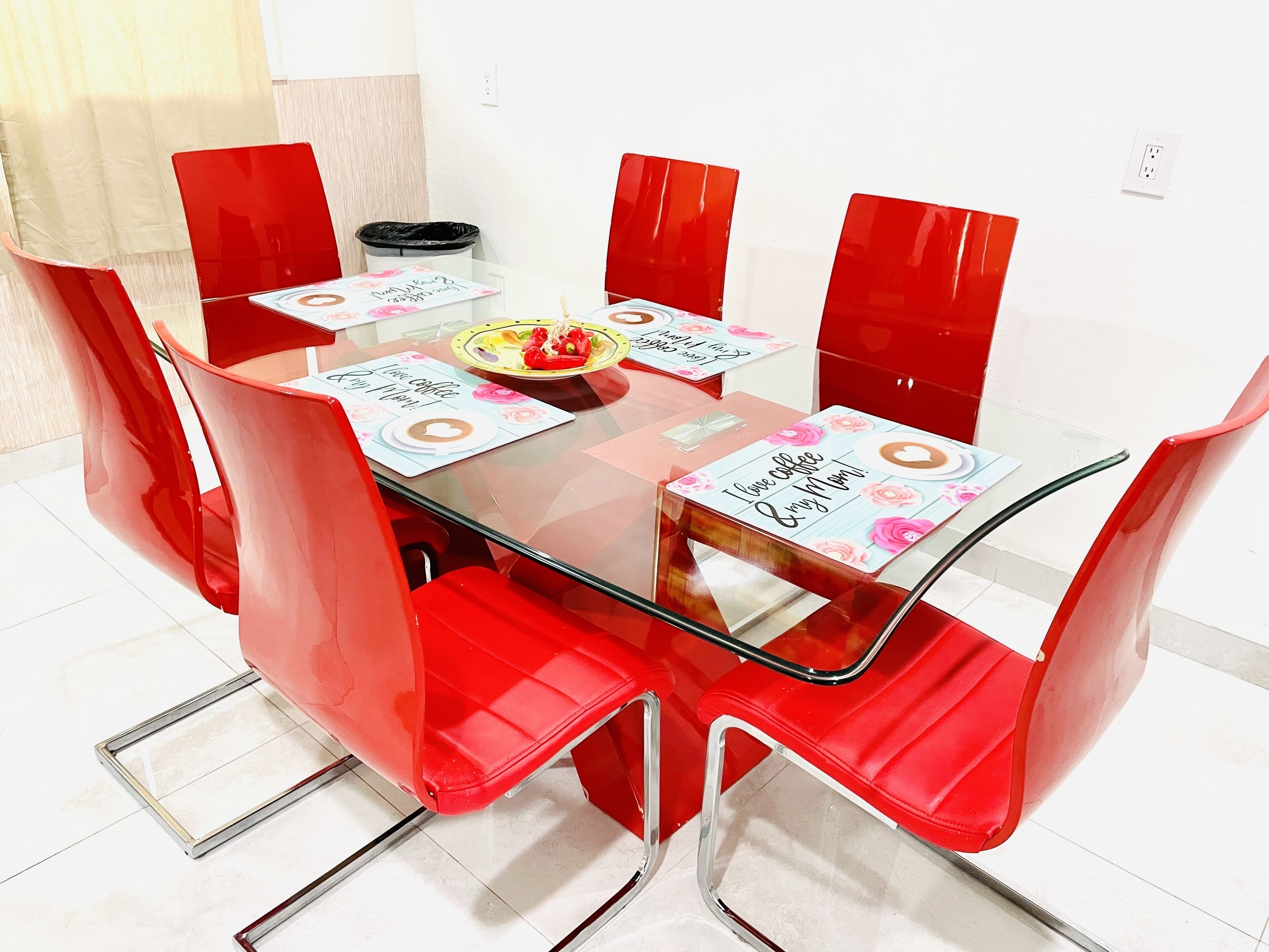 Une table à manger avec des chaises rouges, un plateau en verre transparent et des sets de table aux motifs sur le thème du café.