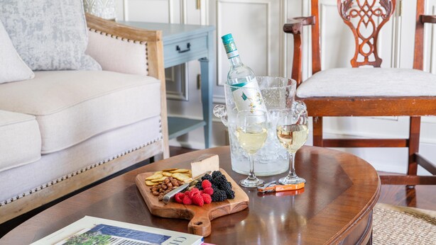 Una mesa de madera con una botella de licor, dos copas de vino blanco, una tabla de madera con aperitivos y una revista.