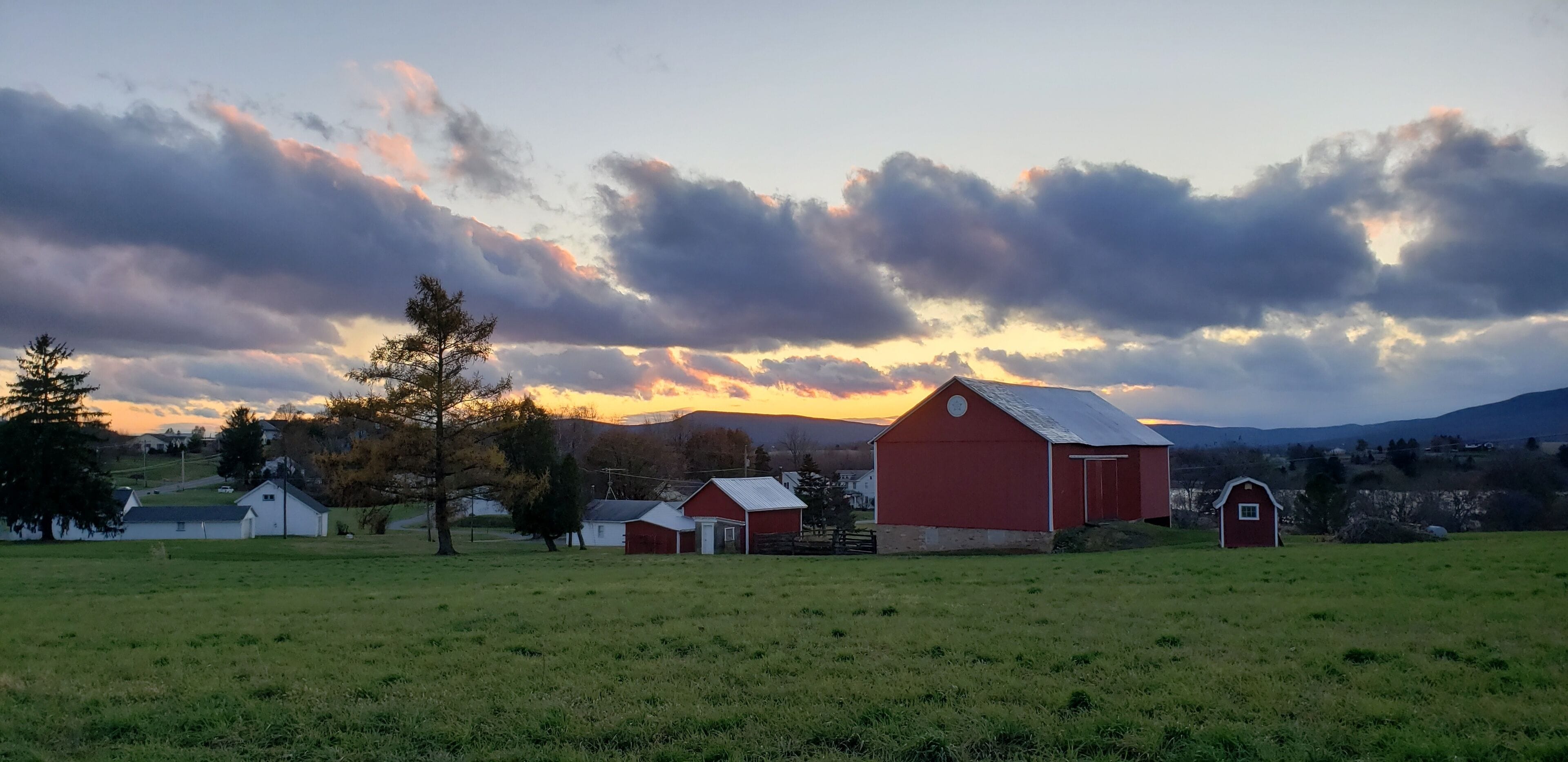 Un paysage rural avec une grange rouge et plusieurs bâtiments plus petits, entourés de champs ouverts et d’arbres, sous un ciel nuageux.