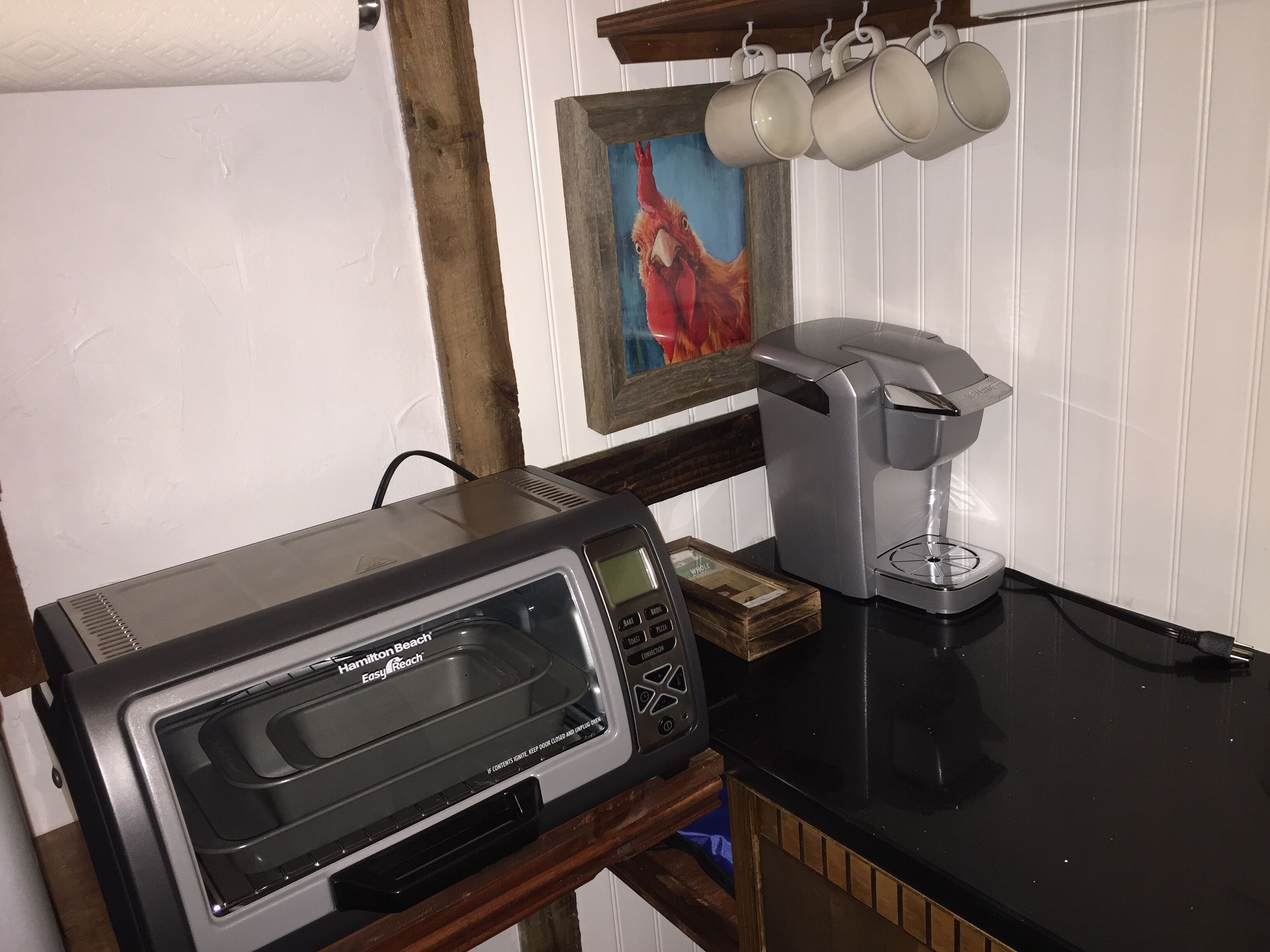 A kitchen with a microwave, coffee maker, and a painting of a rooster on the wall.