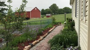 Un camí de maons que condueix a una casa amb un jardí tancat per una tanca, una era vermella i un camp verd al fons.