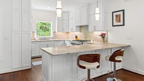A modern kitchen with white cabinetry, a marble island, two bar stools, pendant lights, and a framed picture on the wall. - Fabulous 4 BR/3.5 BA w/ Golf Views! Close to Beach! Amenity access included! No Pets Allowed. (Seabrook Island)