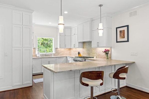 A modern kitchen with white cabinetry, a marble island, two bar stools, pendant lights, and a framed picture on the wall.