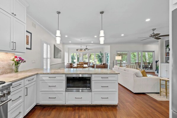 Una cocina moderna con armarios blancos, una isla central con microondas, iluminación colgante y vistas a un área de estar con sofá y mesa de comedor.