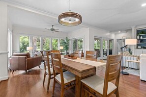 Un comedor con mesa y sillas de madera, ventilador de techo y ventanas amplias con vistas a zonas verdes.