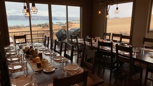 Comedor con mesas dispuestas para cuatro, ventanas amplias con vistas a un lago y montañas, y lámparas colgantes.