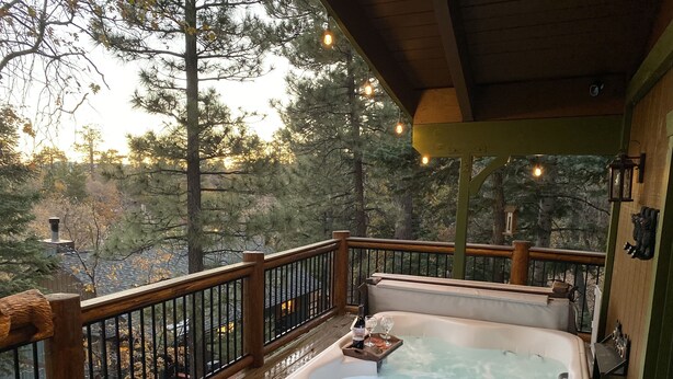 Een hottub op een houten terras met een leuning, omgeven door bomen en uitzicht op een huis op de achtergrond.