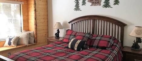 Un dormitorio acogedor con colcha a cuadros, cabecera de madera, mesita de noche con lámpara y ventana con persianas.