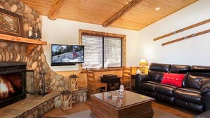 Een gezellige woonkamer met een stenen openhaard, een aan de muur bevestigde televisie, een leren bank en een houten plafond.