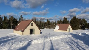 Deux petits bâtiments d’un seul étage, avec des toits bruns et des murs de couleur beige, entourés de neige et d’arbres.