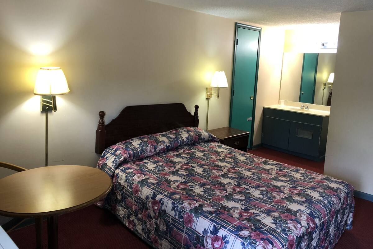 A single bed with a floral comforter, a bedside table, a lamp, a small round table, a mirror, and a bathroom with a sink and cabinet.