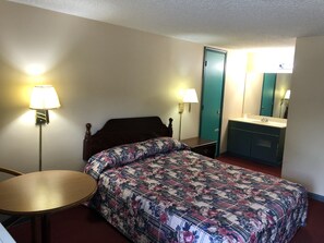 A single bed with a floral comforter, a bedside table, a lamp, a small round table, a mirror, and a bathroom with a sink and cabinet.