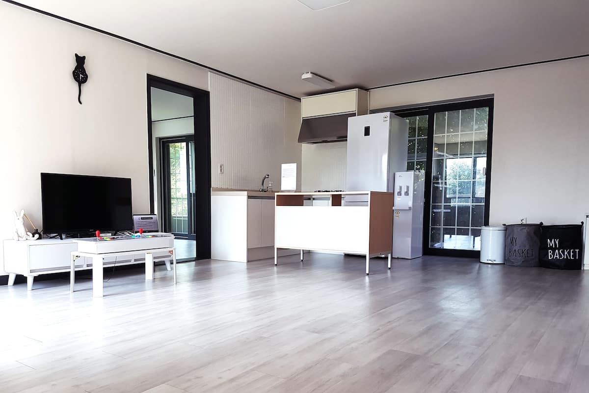 A modern kitchen with a white countertop, a built-in oven, and a refrigerator. There is a dining table with chairs, a TV, and a small table with a laptop and a cup. The room has wooden flooring and large windows.