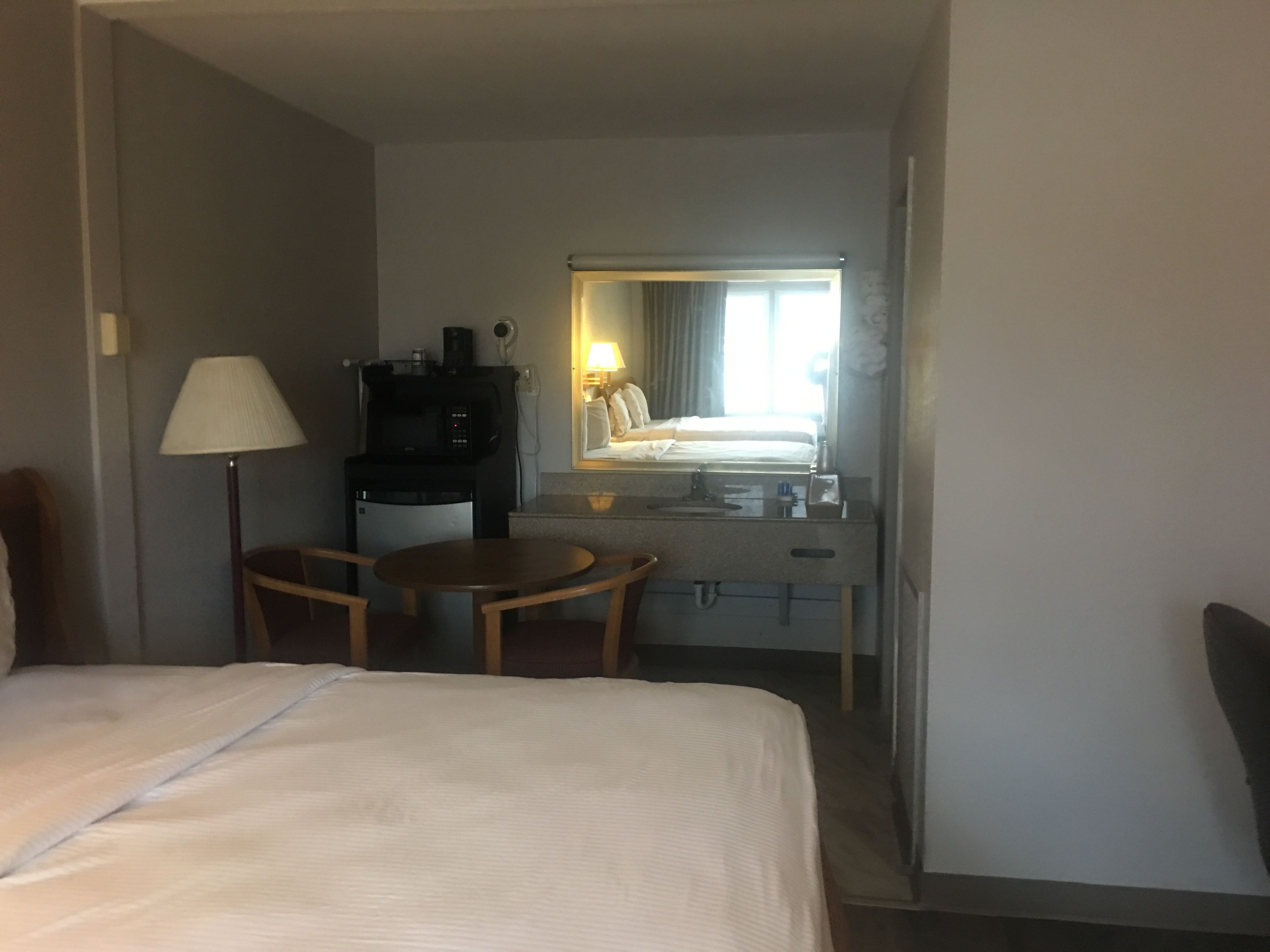 Habitación de hotel con cama, escritorio, silla, lámpara, microondas y espejo.