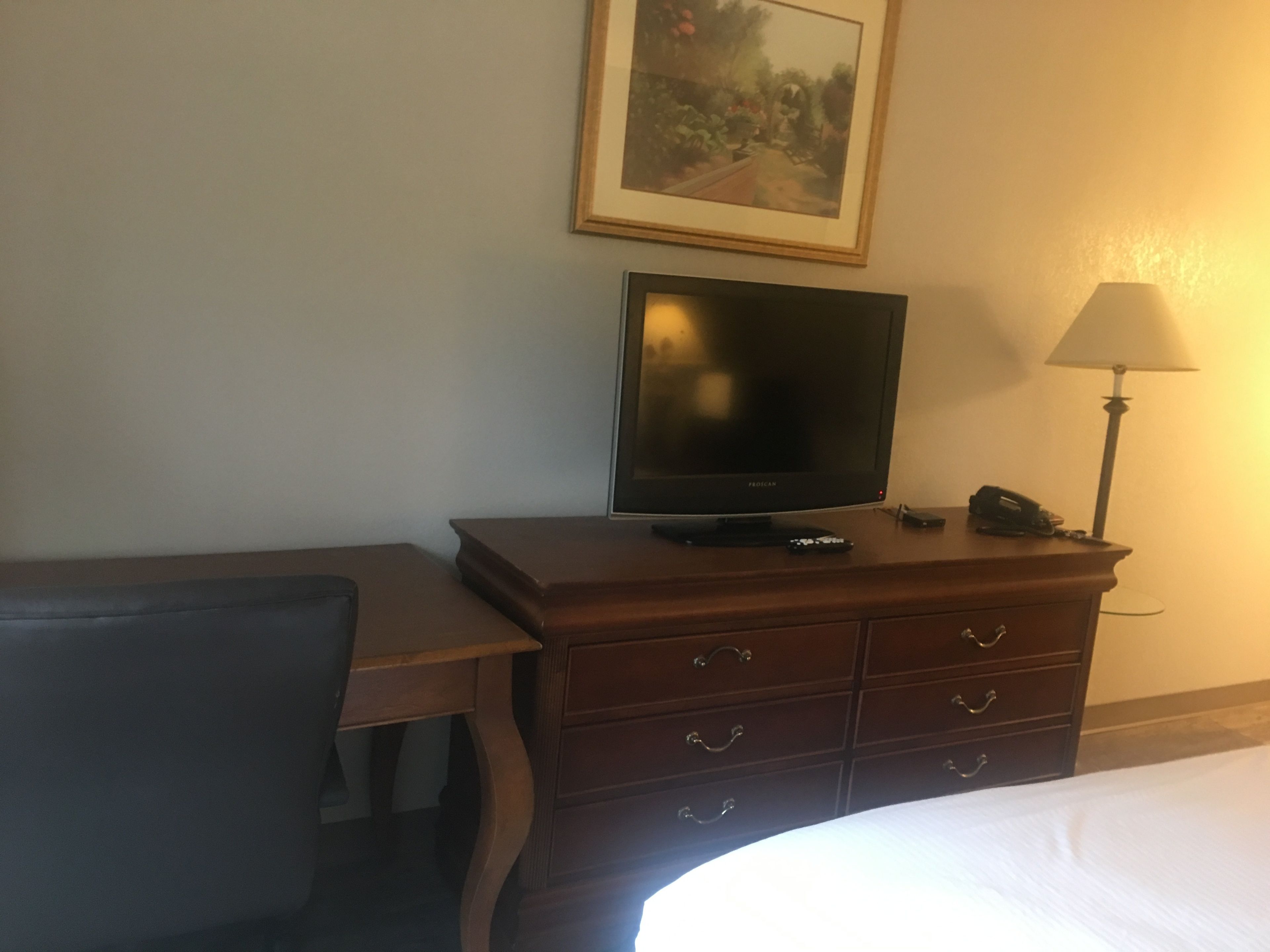 Une chambre d’hôtel avec un lit, une commode en bois avec des tiroirs, un téléviseur posé dessus, un bureau et une lampe.