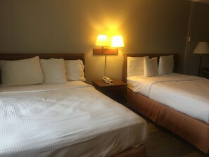 A hotel room with two beds, each with white linens and pillows, a nightstand with a lamp and a telephone, and a wooden headboard.