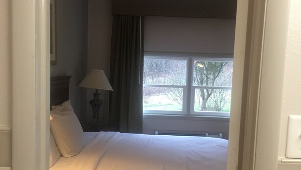 A bedroom with a bed, a nightstand, a lamp, a window with a view of trees, and a door frame. - Relax Inn (Lewisburg)