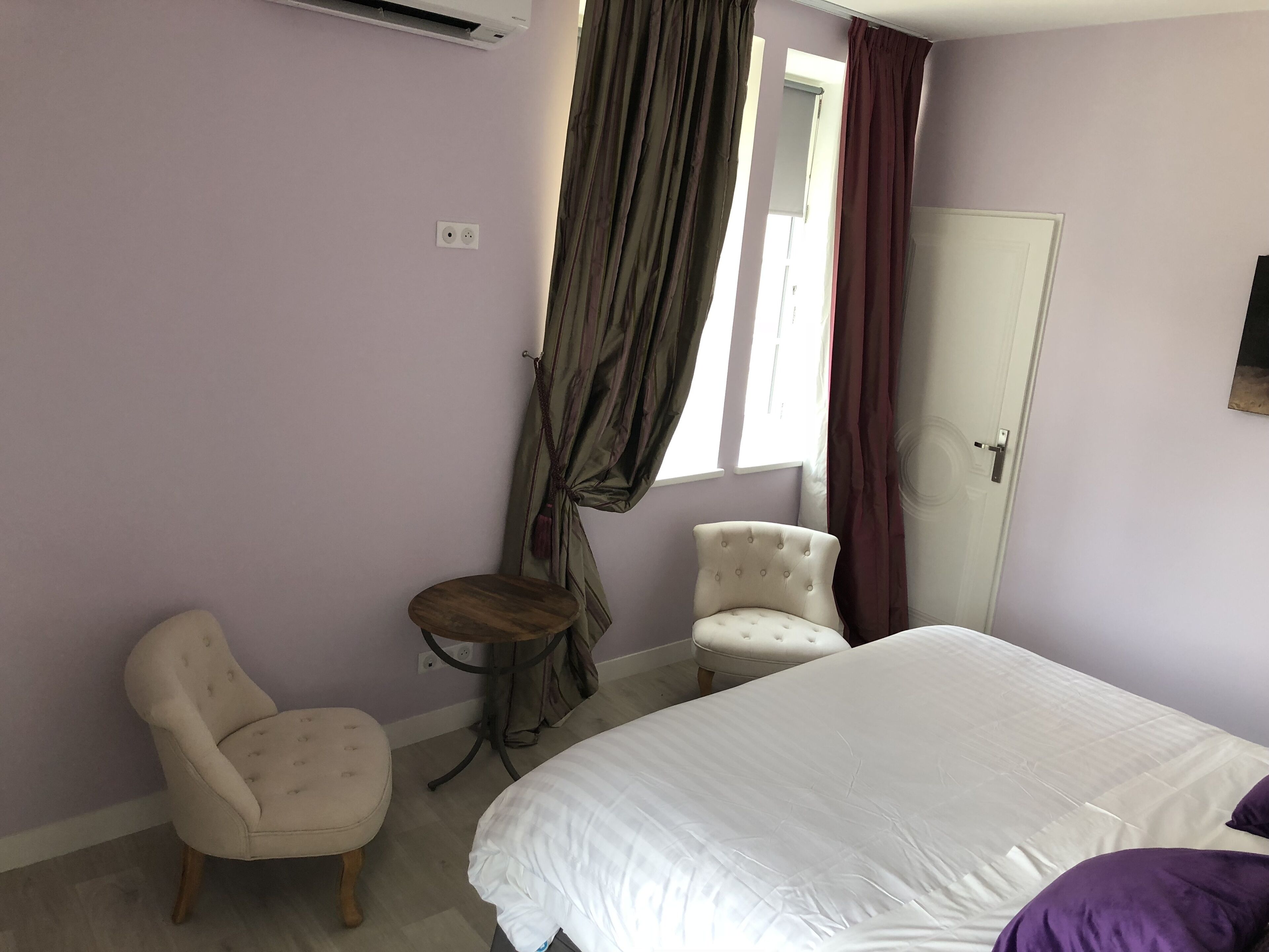 Un dormitorio con cama, dos sillas, una mesita, una ventana con cortinas y una puerta.