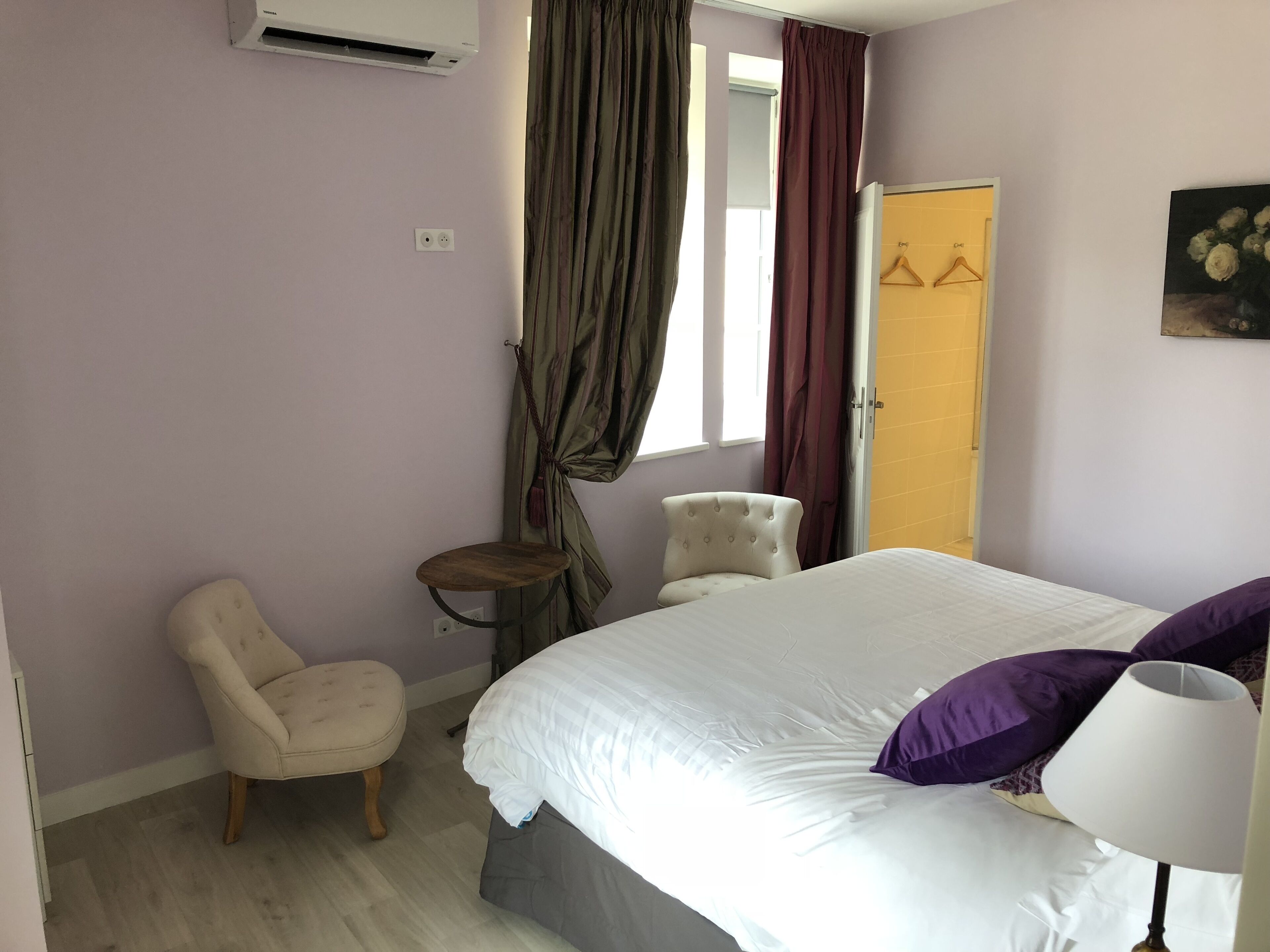 Ein Schlafzimmer mit Bett, Stuhl, kleinem Tisch, Lampe, einem Gemälde und einer Tür.