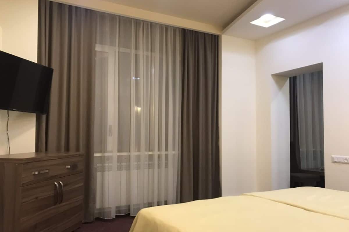 A hotel room with a bed, a wooden dresser, a flat-screen TV, and a large window with sheer curtains.