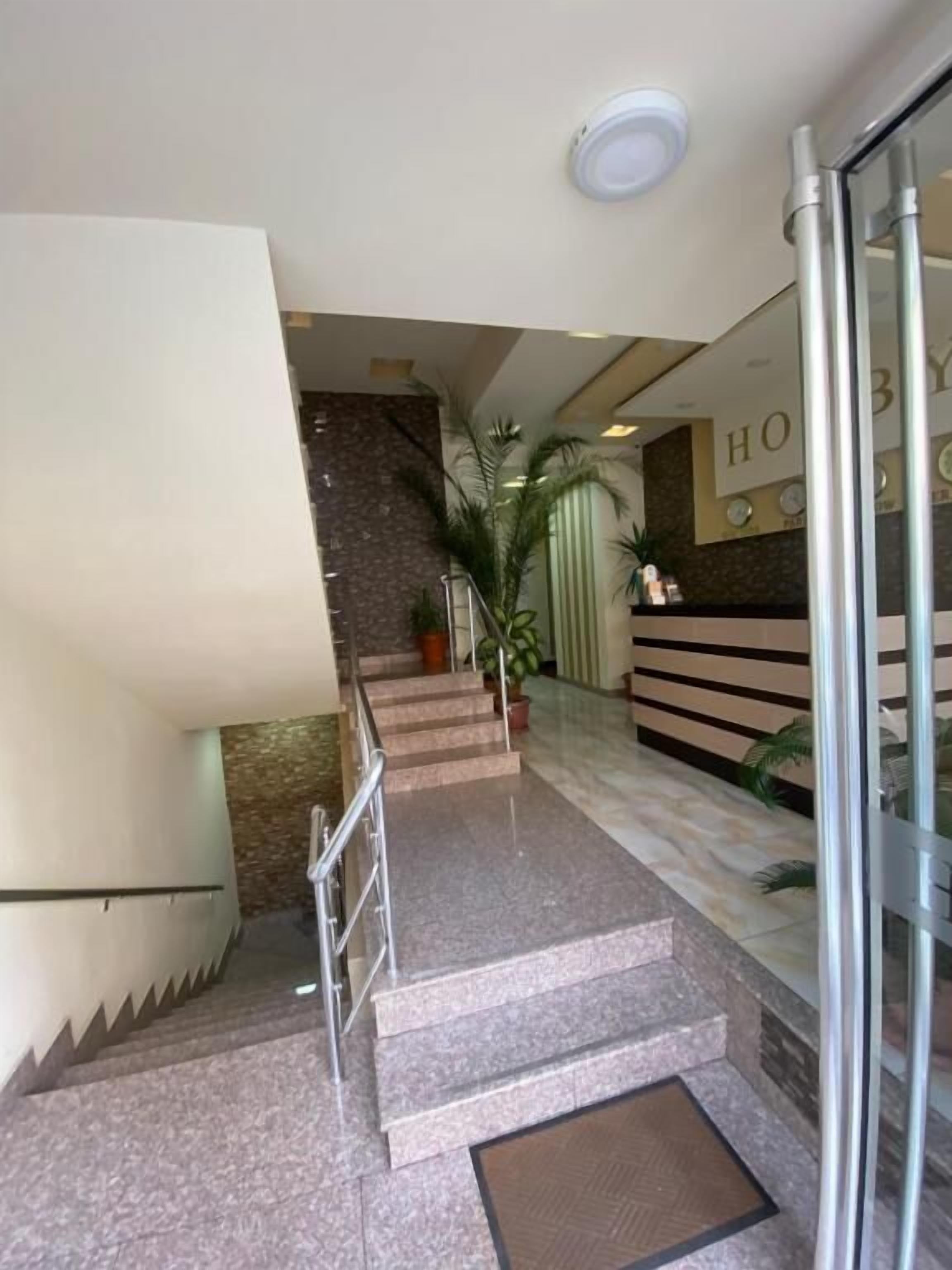A hotel reception area with a reception desk, a staircase leading to an upper floor, and a glass door entrance.