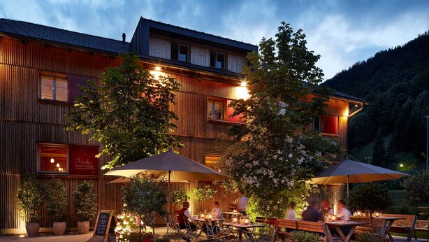 Un área de comedor al aire libre con mesas y sombrillas, rodeada de plantas en macetas y un edificio con revestimiento de madera.