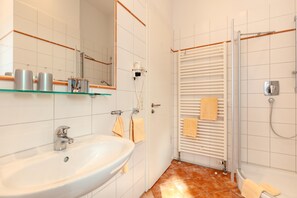 Une salle de bain avec un lavabo blanc, une étagère en verre où sont rangés les articles de toilette, un porte-serviettes et un espace douche.