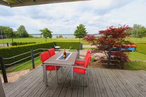 Una terraza de madera con una mesa y sillas rojas, con vistas a un jardín ajardinado y una masa de agua.