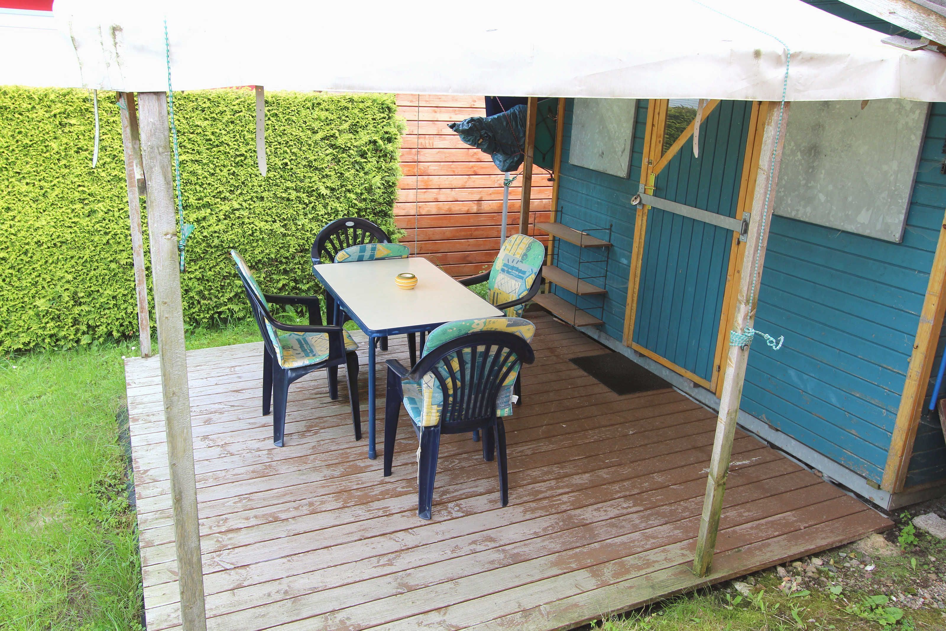 Una terraza de madera con mesa y sillas, un área cubierta y un edificio azul con una puerta.