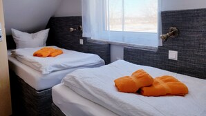 Dos camas individuales con sábanas blancas y toallas naranjas, una ventana con cortinas translúcidas y una lámpara fijada a la pared.