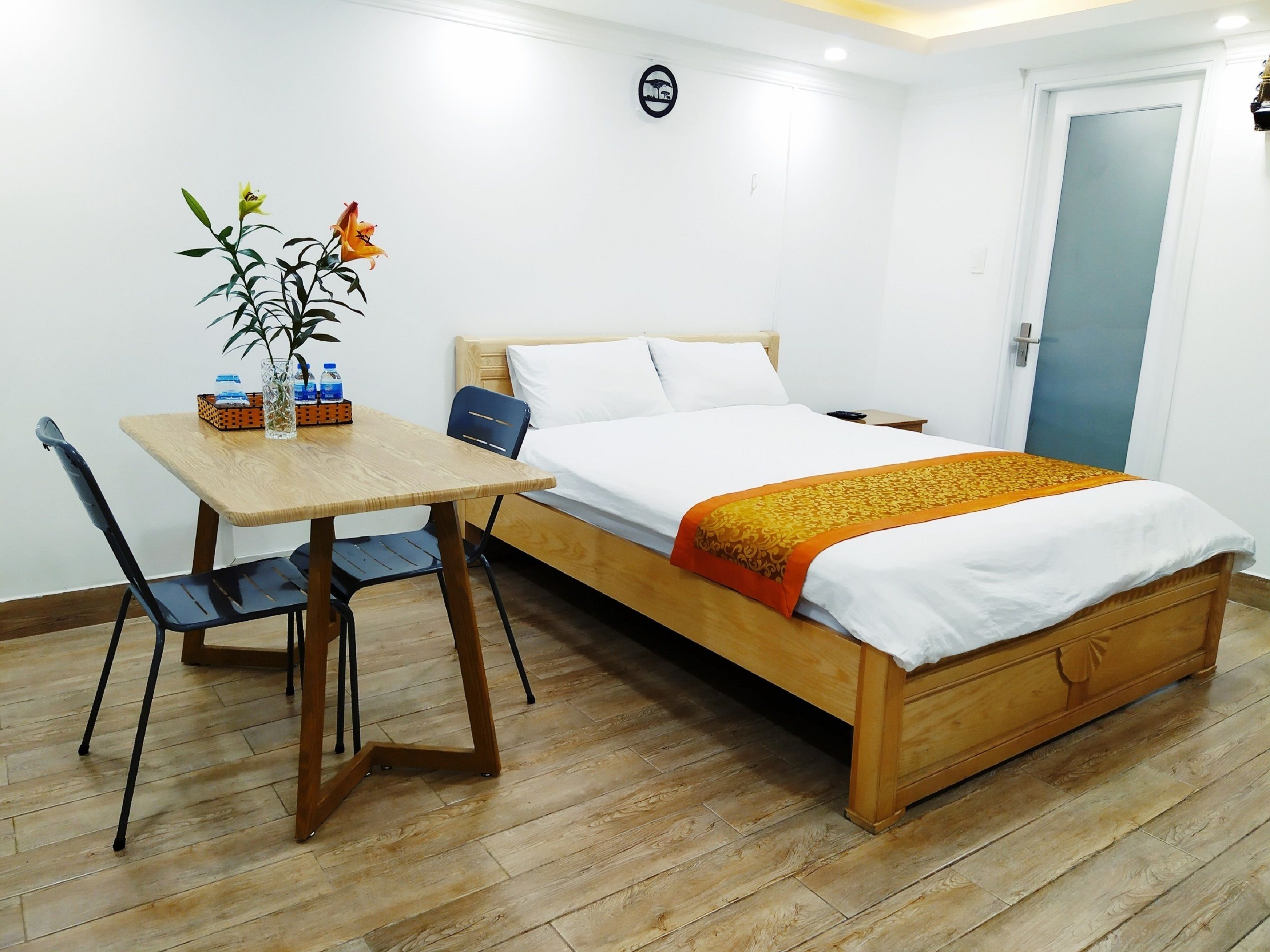 Een hotelkamer met een houten bed, een kleine houten tafel met stoelen, een vaas met bloemen en een deur.