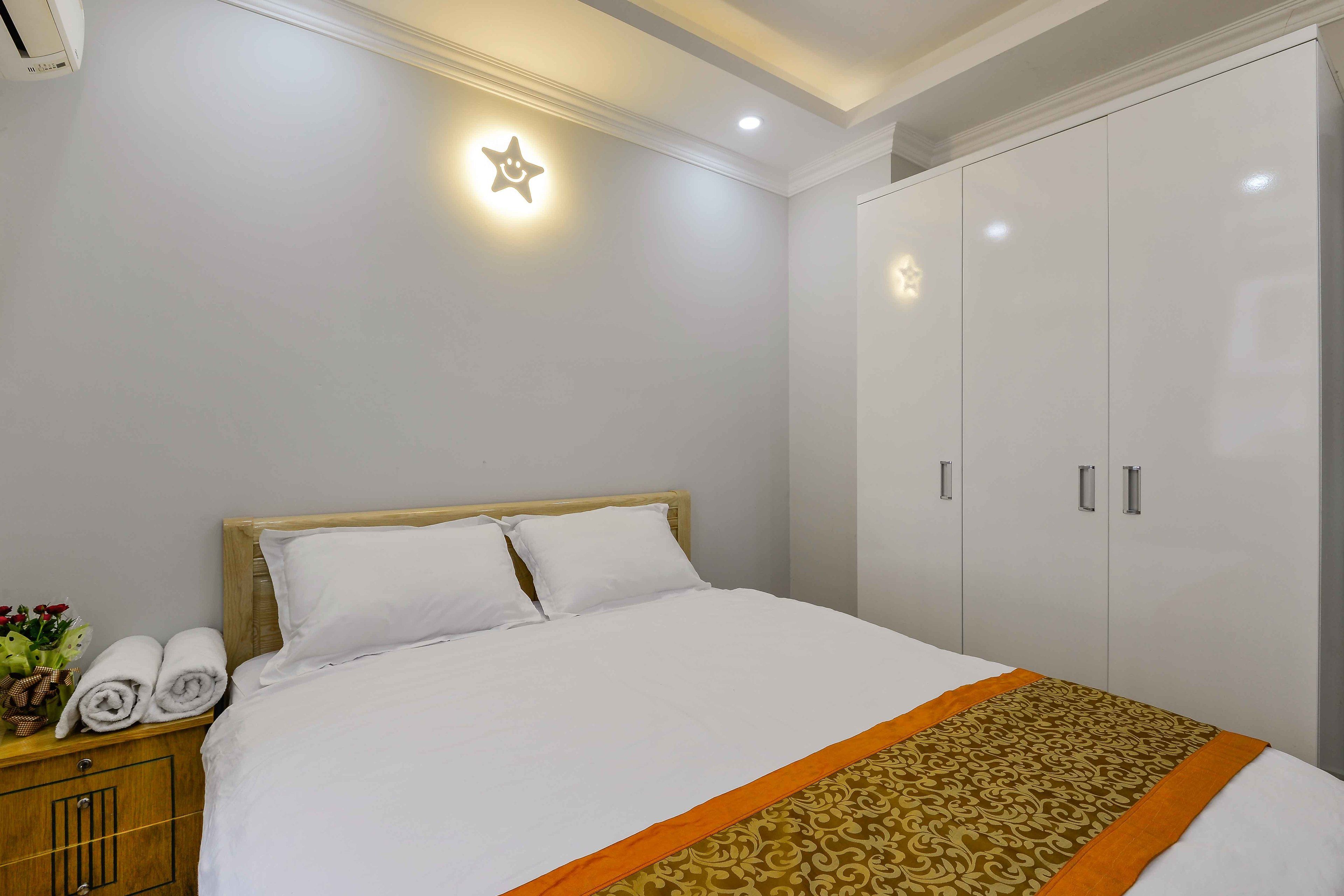 A neatly made bed with white linens and an orange decorative border, a wooden nightstand with rolled towels, and a wardrobe with silver handles.