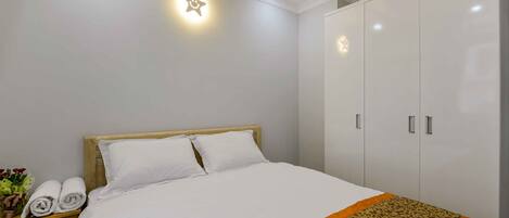 A neatly made bed with white linens and an orange decorative border, a wooden nightstand with rolled towels, and a wardrobe with silver handles.