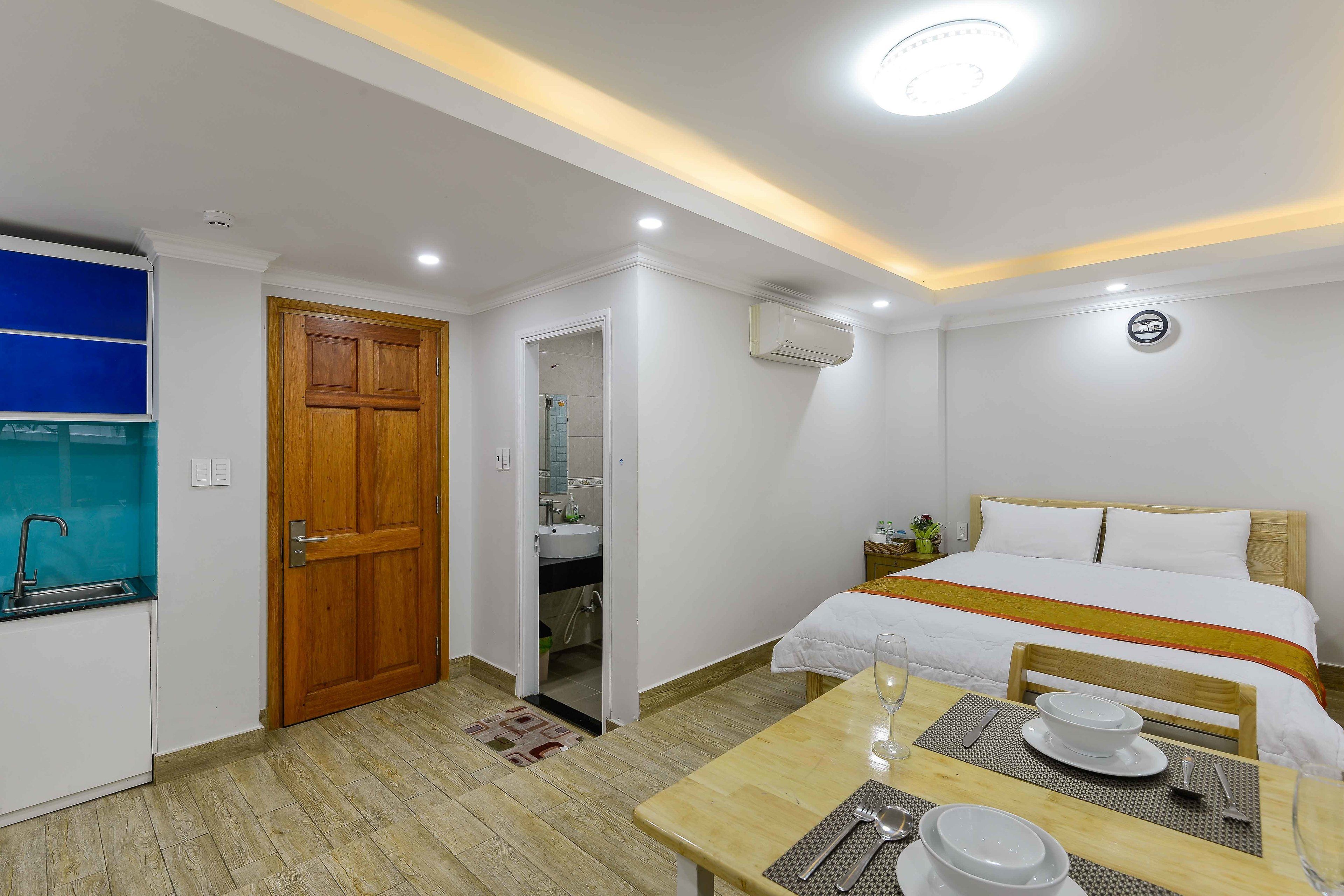 A hotel room with a bed, a wooden door, a bathroom, a sink, a table with a glass, and a small plant.