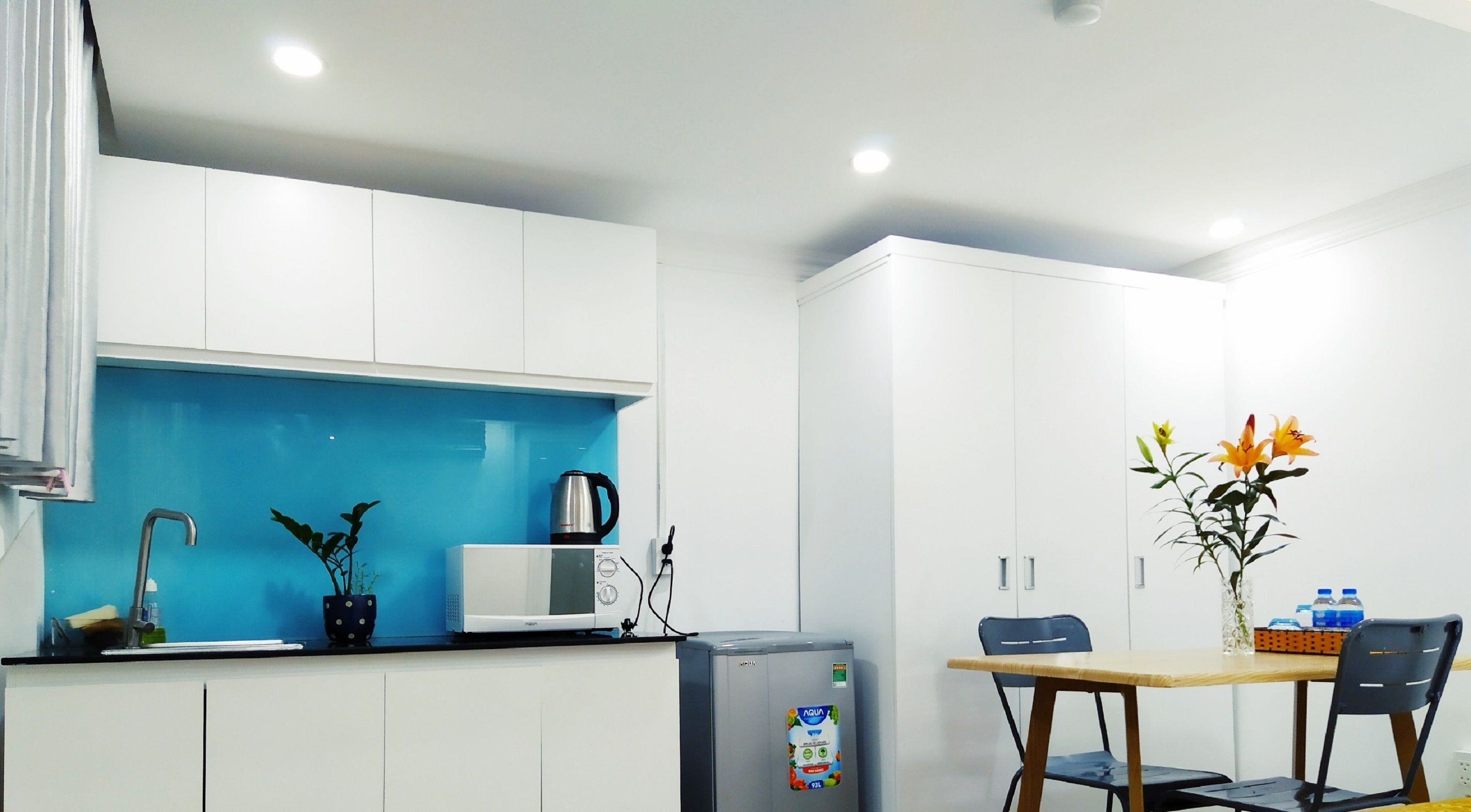 A compact kitchen with white cabinets, a blue backsplash, a microwave, a kettle, and a refrigerator.