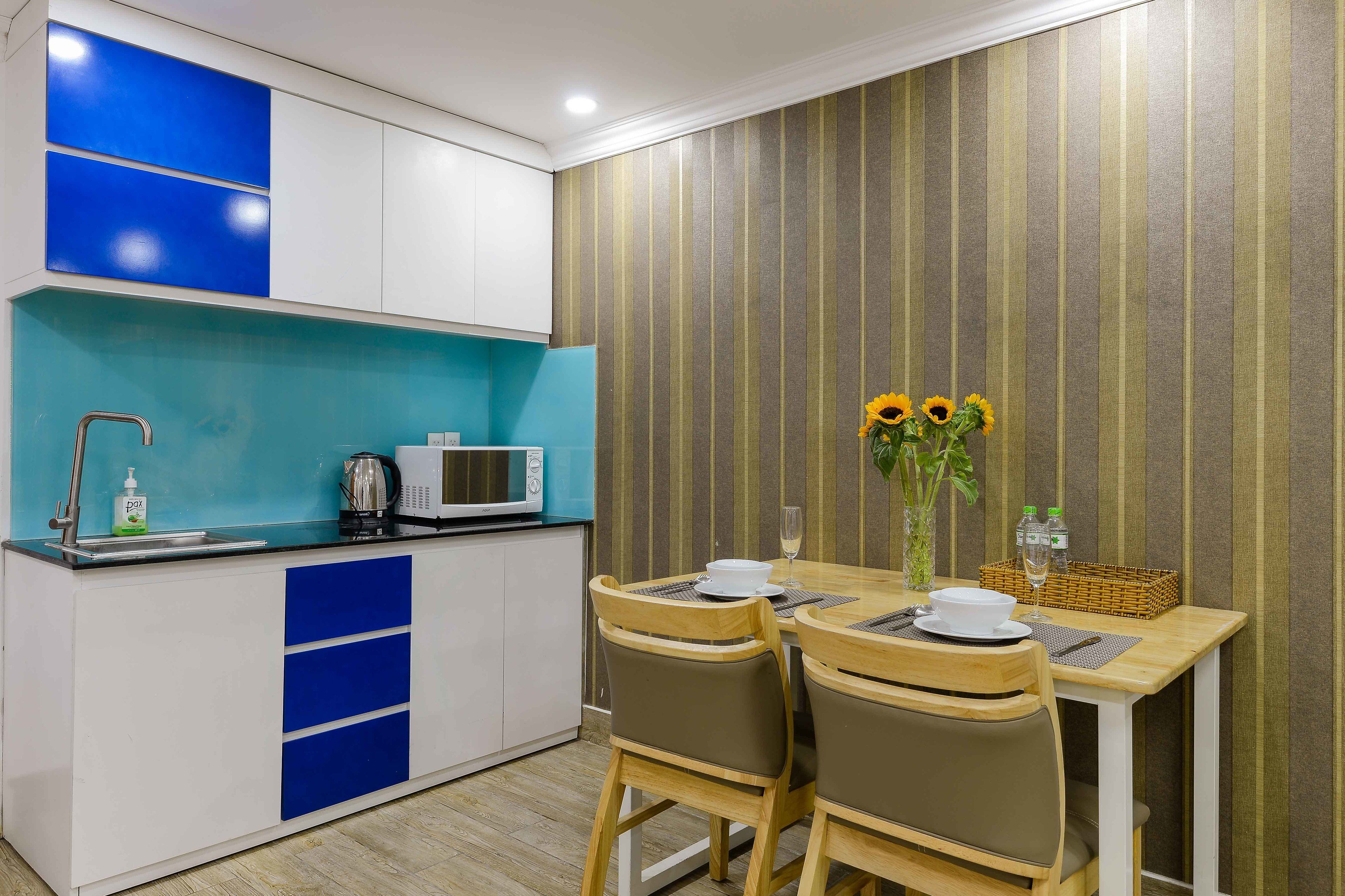 A compact kitchen with a microwave, sink, and a teal backsplash. There is a dining area with two chairs and a table, and a vase with sunflowers.