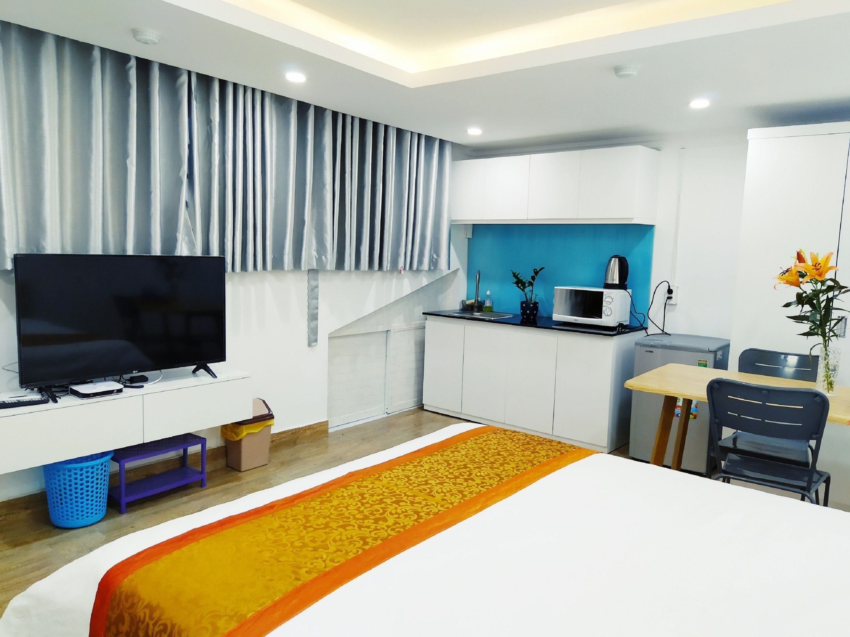 Habitación de hotel moderna con televisión de pantalla plana, una kitchenette pequeña, zona de comedor y cama con una colcha decorativa.
