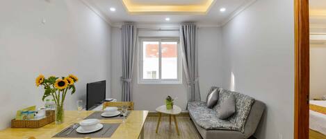 A compact living space with a dining area, a sofa, a small table with a vase of sunflowers, and a window with curtains.