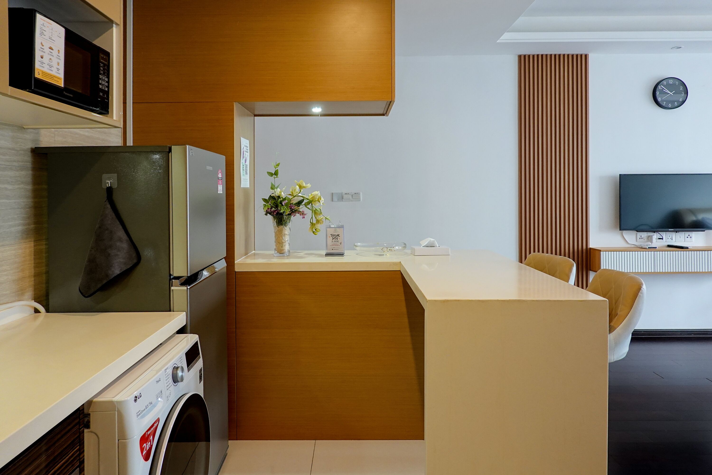 A compact kitchen with a washing machine, a refrigerator, a microwave, and a dining area with a TV and a clock on the wall.
