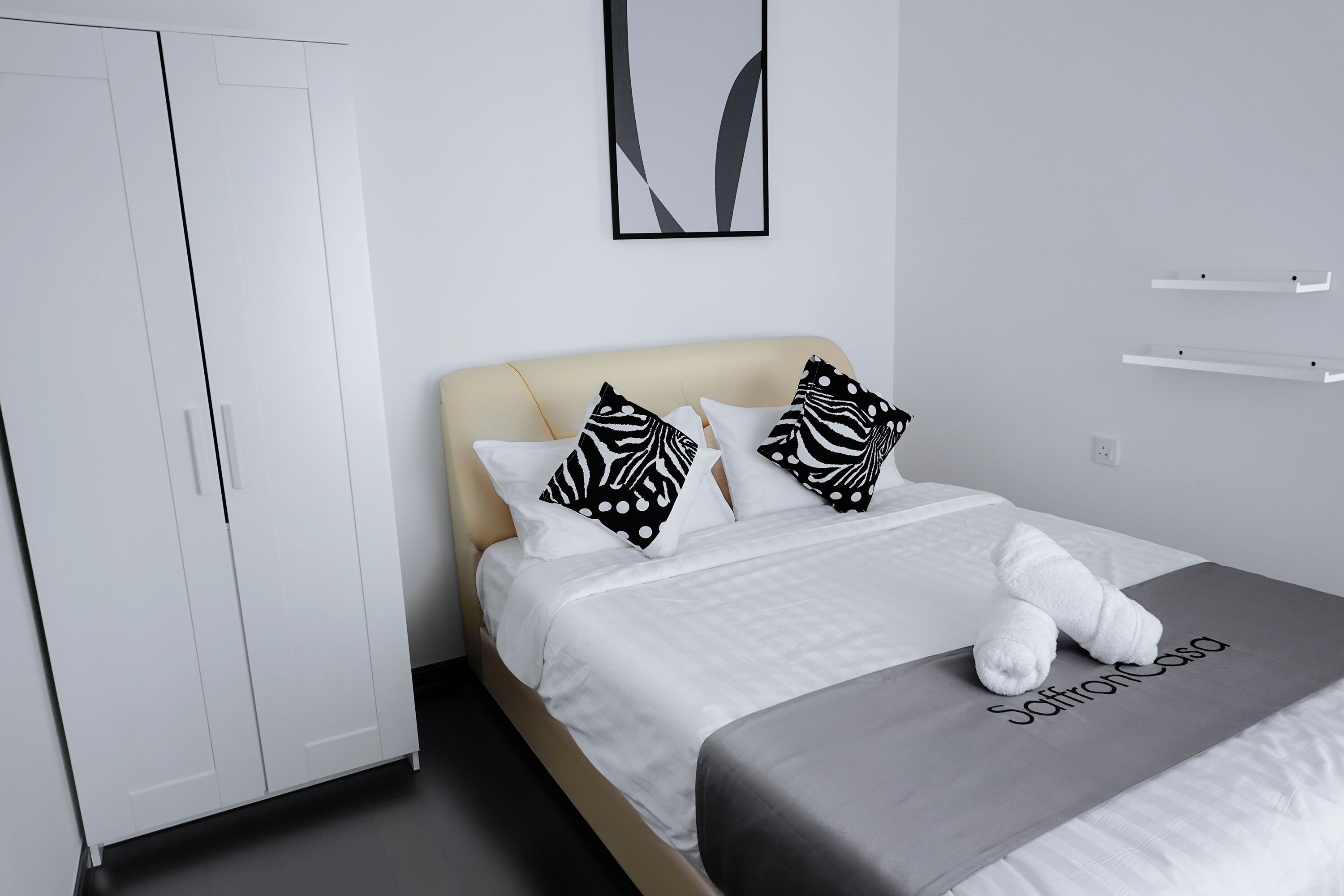 Una cama bien hecha con ropa de cama blanca y almohadas estampadas en blanco y negro, una manta gris con el texto "SaffronCasa" y un armario blanco.
