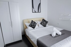 A neatly made bed with white bedding and black and white patterned pillows, a grey blanket with "SaffronCasa" text, and a white wardrobe.