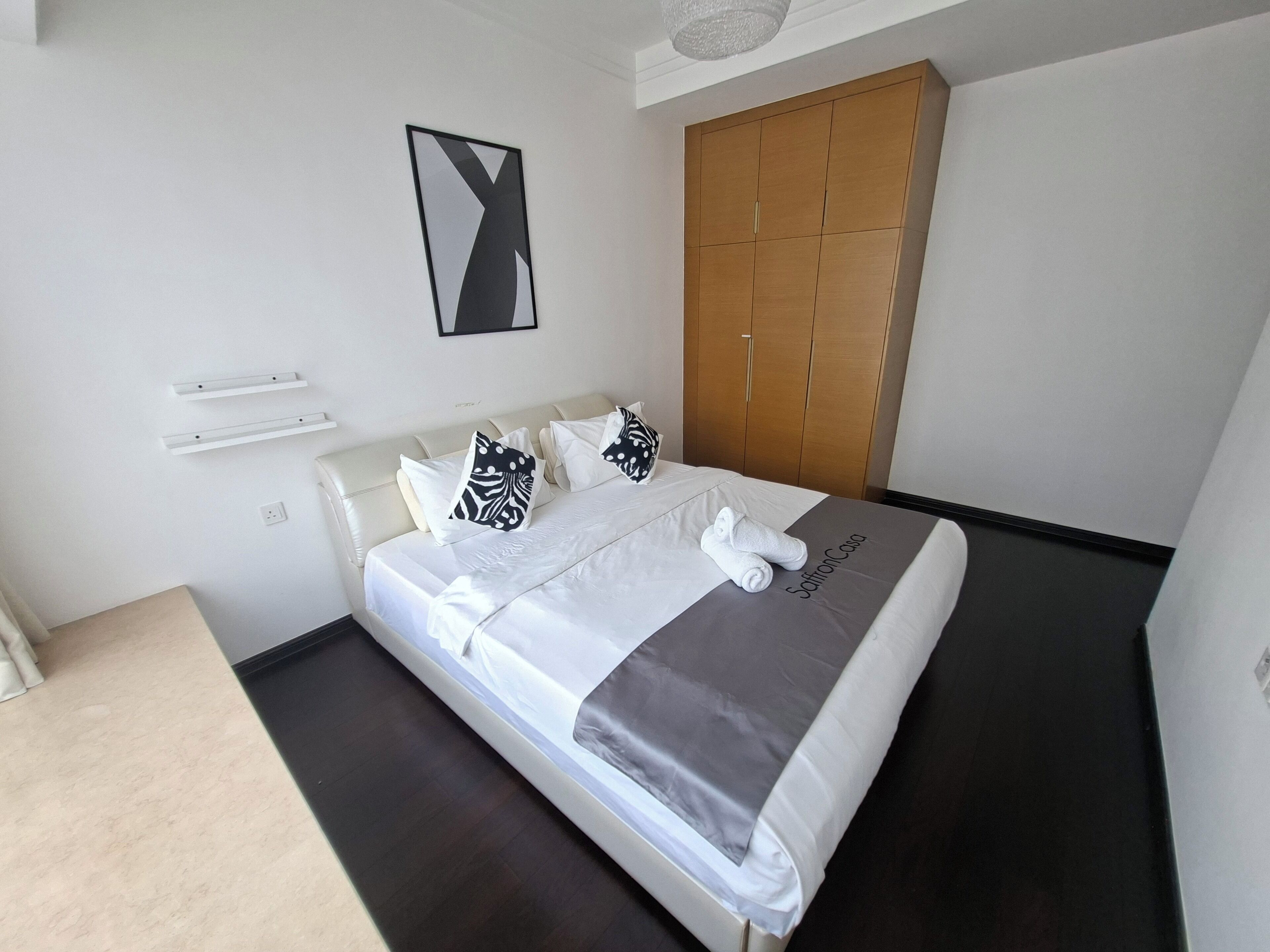 Un lit bien fait, recouvert d’une couverture grise, avec des oreillers blancs et une serviette. On aperçoit une armoire en bois et un tableau encadré au mur.