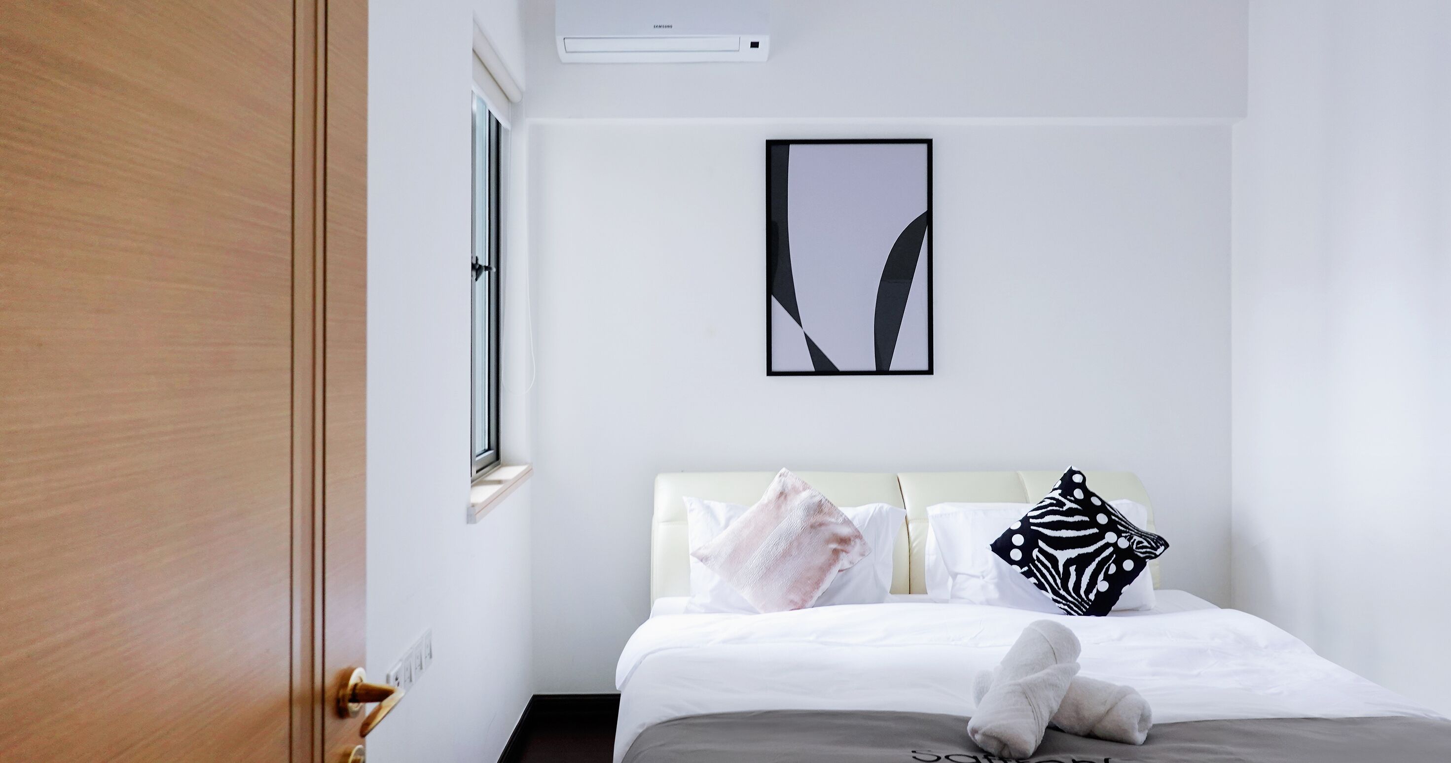 A hotel room with a bed, a window, a wall-mounted air conditioner, and a framed abstract artwork.