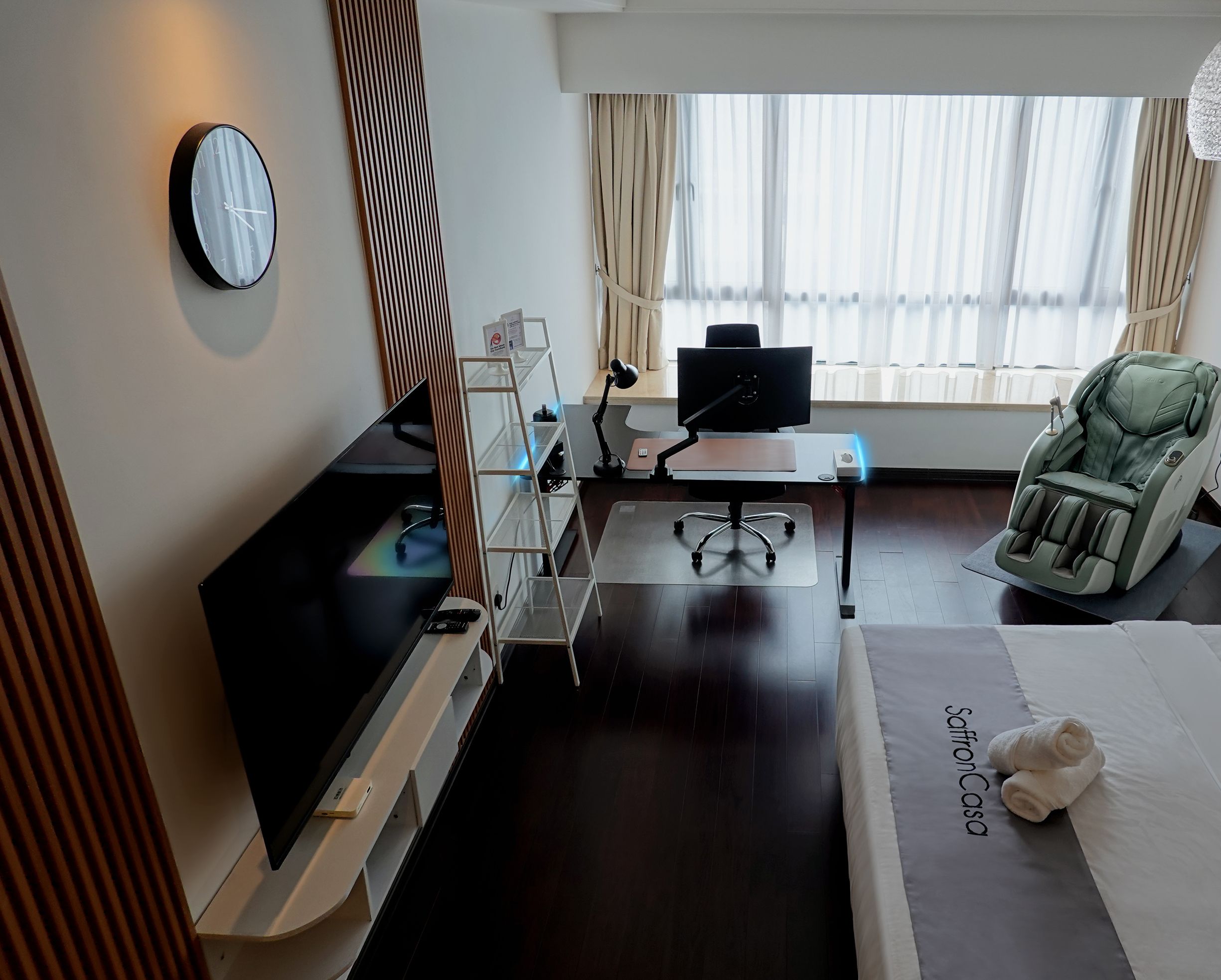 Une chambre d’hôtel équipée d’un lit, d’un bureau avec un ordinateur, d’un fauteuil massant, d’une télévision et d’une horloge murale.