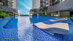 A swimming pool with a tiled floor and lounge chairs, surrounded by tall buildings and palm trees. - R&F Princess Cove JB By SaffronCasa JB (Johor Bahru)