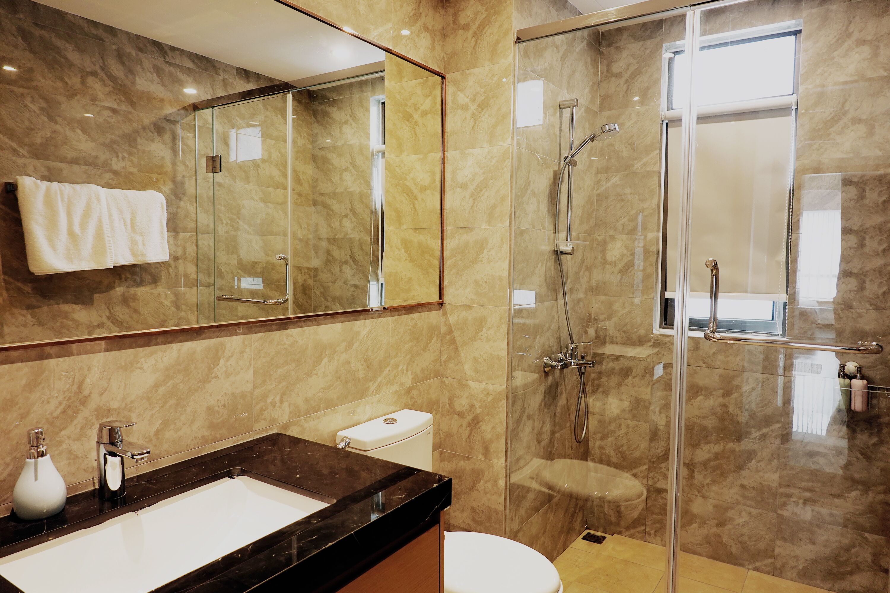 Ein modernes Badezimmer mit einer gläsernen Duschkabine, einer Marmorwand und einem großen Spiegel sowie einer weißen Toilette.