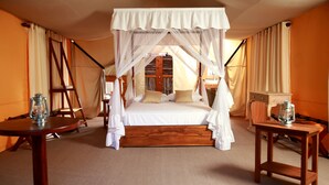 Una cama con dosel sobre cuatro postes, con estructura de madera y ropa de cama blanca, una pequeña mesa de madera con un farol y una silla con cojín.
