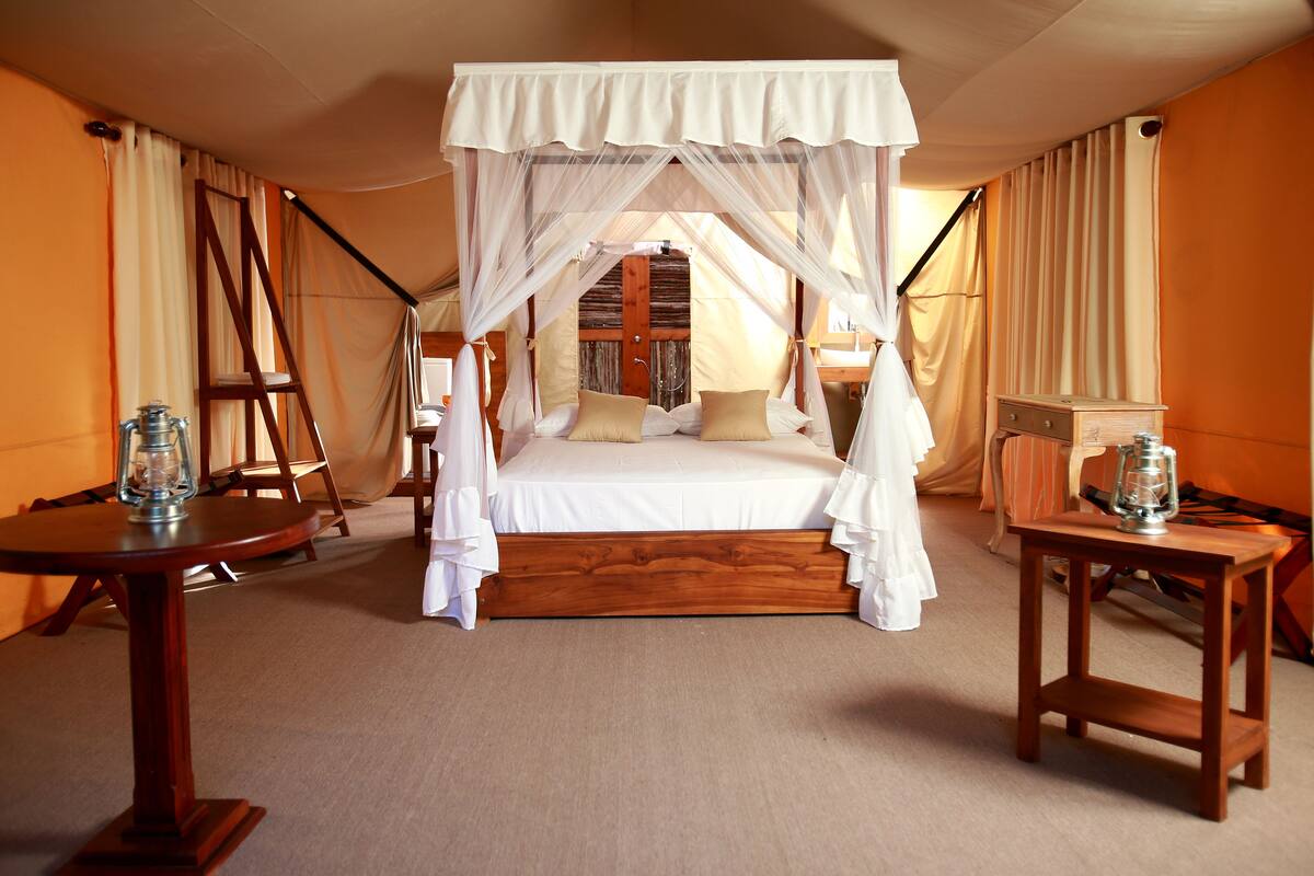 Un lit à baldaquin à quatre poteaux, avec une structure en bois et du linge de lit blanc, une petite table en bois sur laquelle est posée une lanterne, et une chaise avec un coussin.