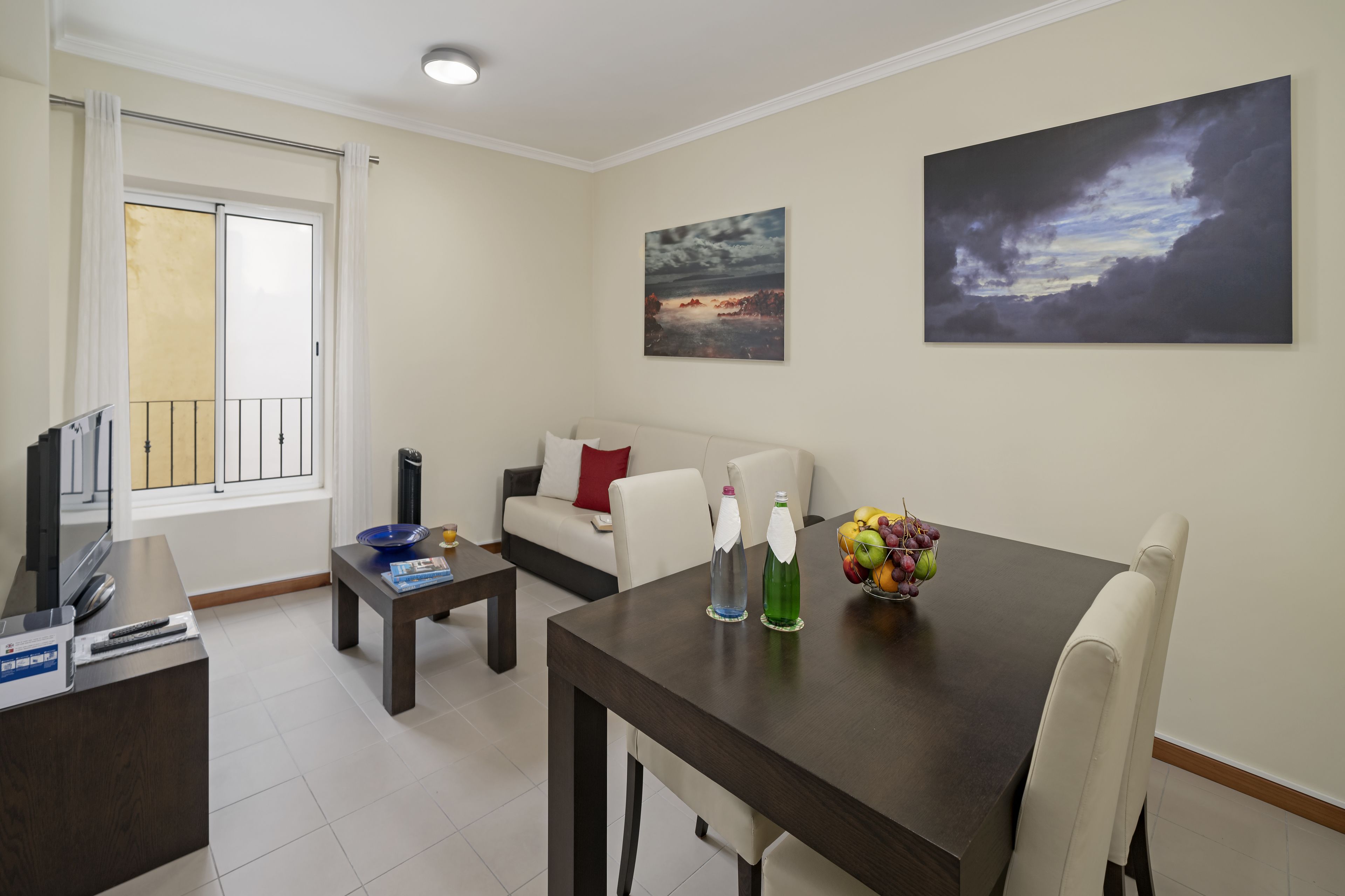 A living room with a sofa, a dining table with chairs, a TV, and a window with curtains.