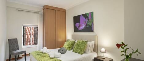 Un dormitorio ordenado con una cama, una silla, una mesita de noche y un florero con una flor.